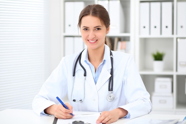 Young brunette female doctor sitting at the table and working at hospital office.  Health care, insurance and help concept. Physician ready to examine patient