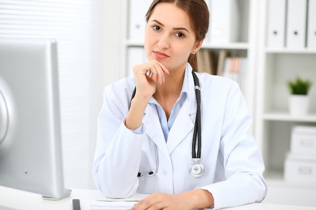 Young brunette female doctor sitting at the table and working at hospital office.  Health care, insurance and help concept. Physician ready to examine patient