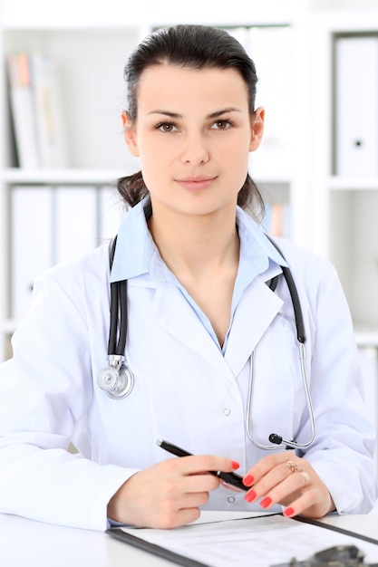 Young brunette female doctor sitting at the table and working at hospital office.  Health care, insurance and help concept. Physician ready to examine patient