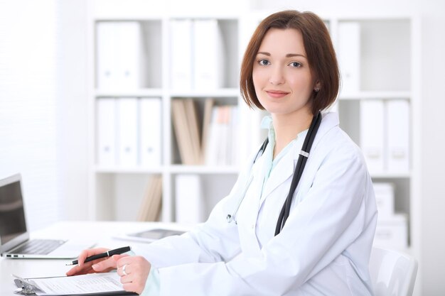 Young brunette female doctor sitting at the table and working at hospital office.  Health care, insurance and help concept. Physician ready to examine patient