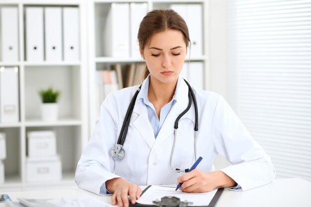 Young brunette female doctor sitting at the table and working at hospital office.  Health care, insurance and help concept. Physician ready to examine patient