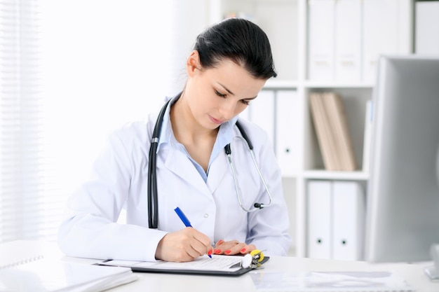 Young brunette female doctor sitting with clipboard near window in hospital and filling up medical history form