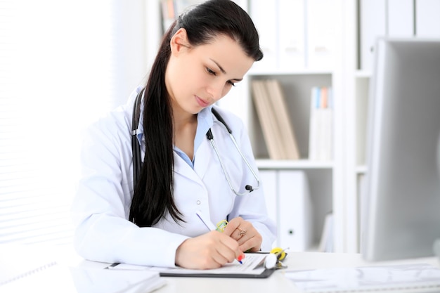 Young brunette female doctor sitting with clipboard near window in hospital and filling up medical history form