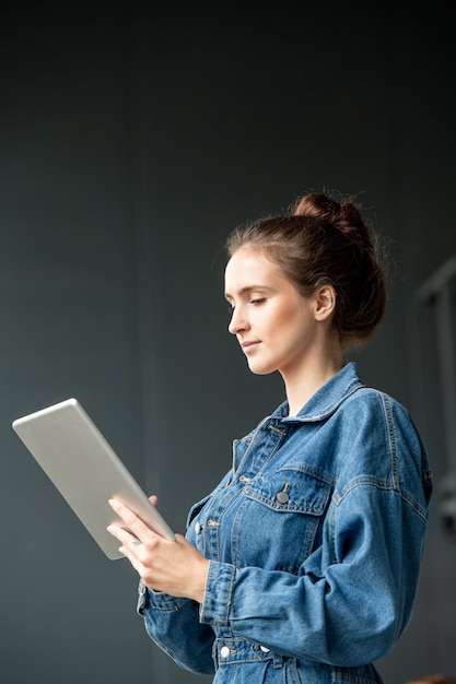 Young brunette female office worker in denim casualwear using digital tablet while scrolling through online videos or information