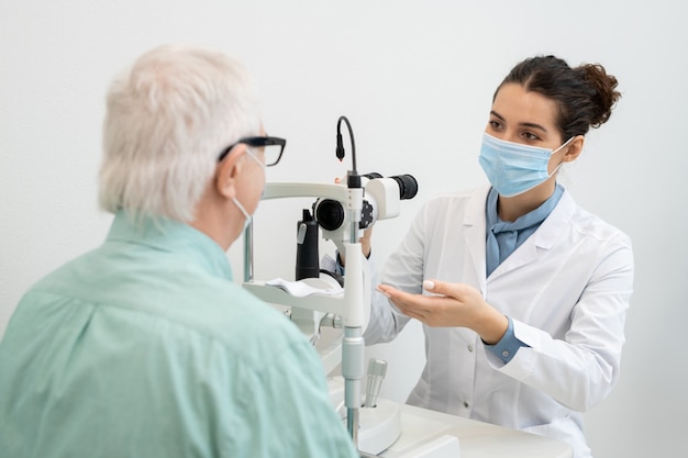 彼女の前に座っている年配の男性患者の視力をテストするために行くホワイトコートと保護マスクの若いブルネットの女性眼科医