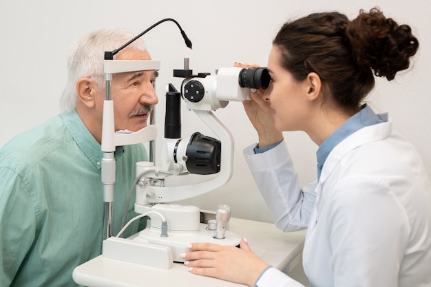 백의를 입은 젊은 브루네트 여성 안과 의사는 노인 남성 환자의 시력을 검사하는 동안 진료소에서 의료 장비를 사용합니다.