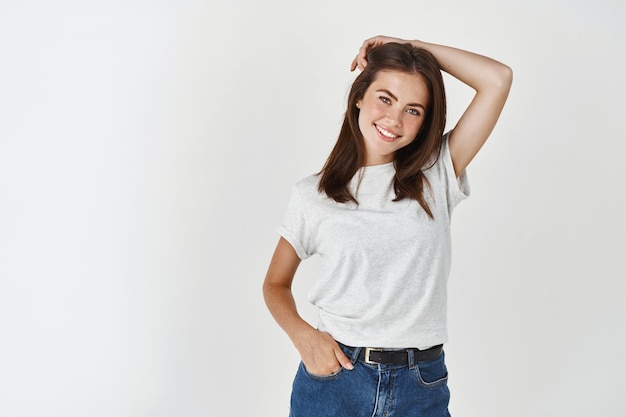 Young brunette female posing in casual white t-shirt against studio wall, smiling and looking happy at at front