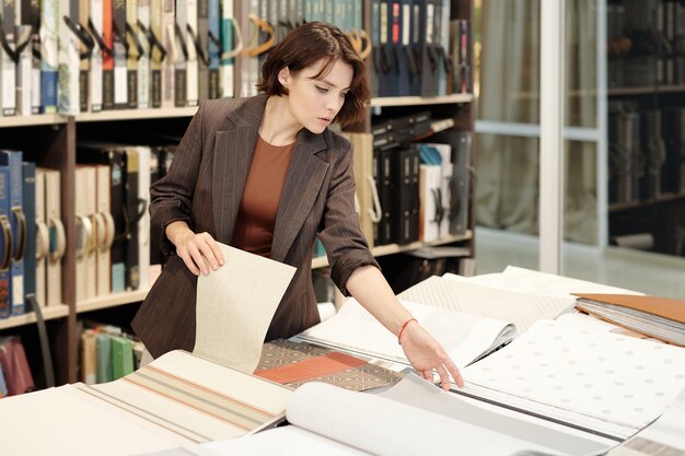 Young brunette female shop assistant or manager of studio of interior design looking through samples of new wallpapers by large table