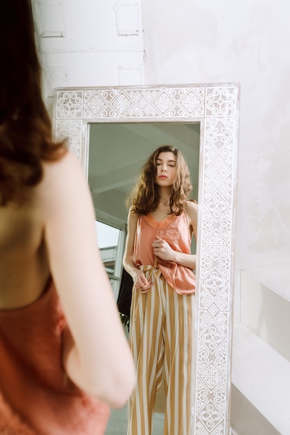 Photo young brunette female in top and striped pants looking at herself in the mirror