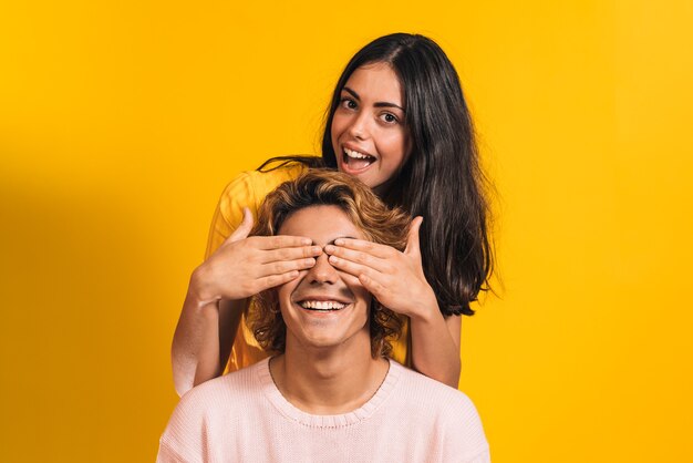 Young brunette girl covering her eyes to a young blond boy on a yellow background, playing