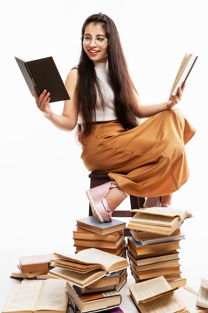 A young brunette girl in glasses with long hair is sitting on a pile of books and reading. Education and training. White background. Vertical.