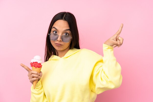 Young brunette girl holding a cornet ice cream over isolated pink background pointing finger to the laterals and happy