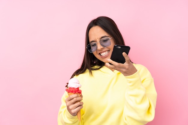 Young brunette girl holding a cornet ice cream over isolated pink background with phone in victory position