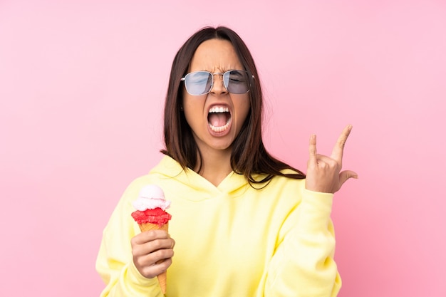 Young brunette girl holding a cornet ice cream over isolated pink wall making rock gesture