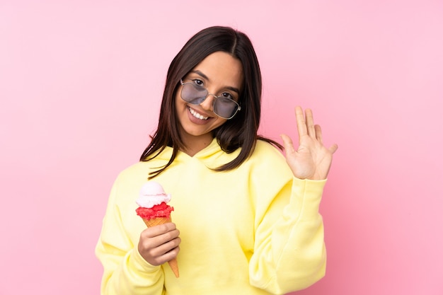 Young brunette girl holding a cornet ice cream over isolated pink wall saluting with hand with happy expression