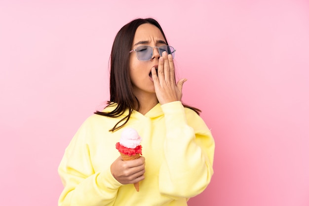 Young brunette girl holding a cornet ice cream over isolated pink wall yawning and covering wide open mouth with hand