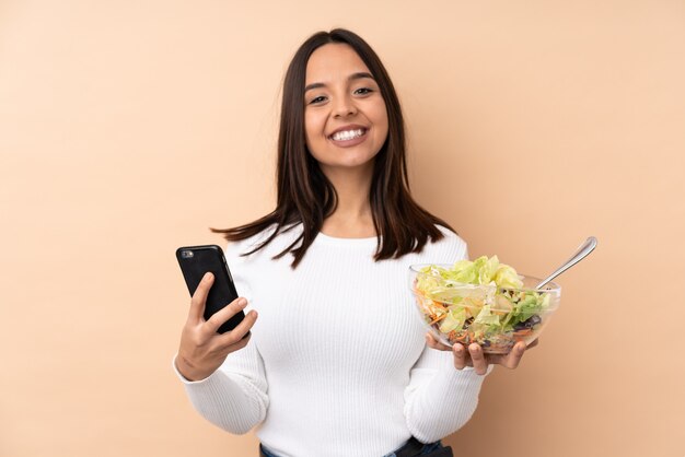 Young brunette girl holding a salad over isolated wall holding coffee to take away and a mobile