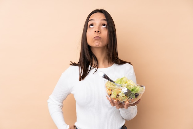 Young brunette girl holding a salad over isolated wall and looking up