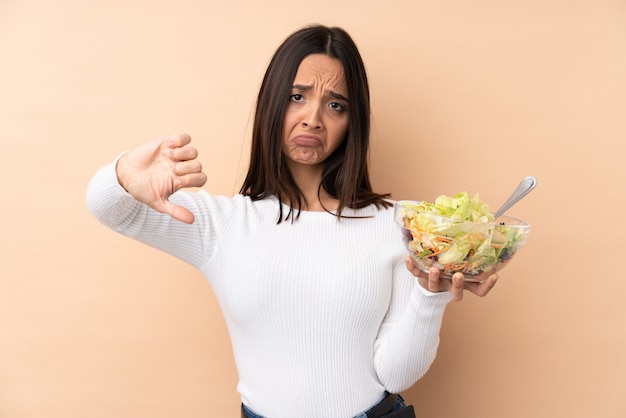 Photo young brunette girl holding a salad over isolated wall showing thumb down