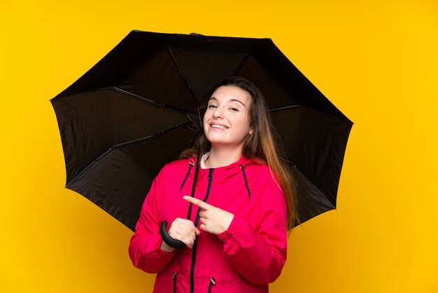 Young brunette girl holding an umbrella over isolated yellow surprised and pointing side