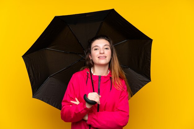 Young brunette girl holding an umbrella over isolated yellow wall smiling a lot