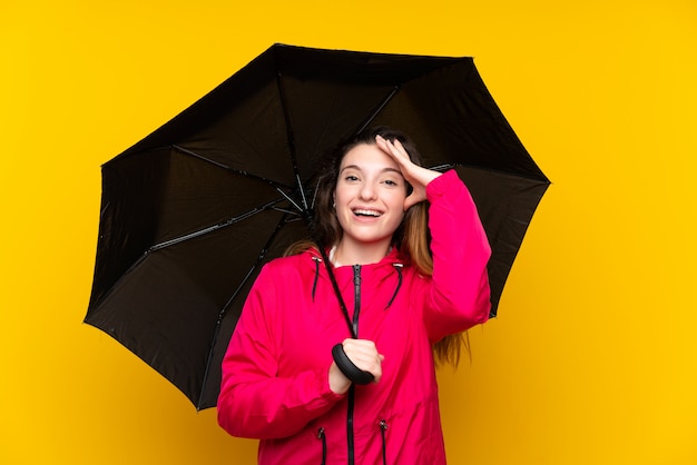 Young brunette girl holding an umbrella over isolated yellow wall with surprise and shocked facial expression