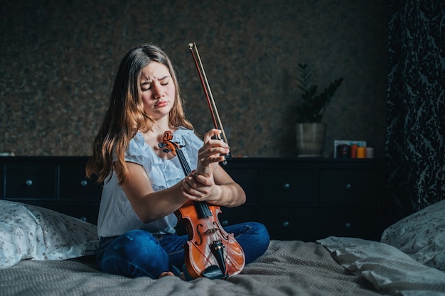 A young brunette girl injured her wrist while playing the violin. A girl sits on a bed in her room and holds a sore wrist with her hand.