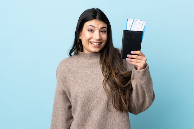 Молодая девушка брюнет над изолированным голубым счастливым в каникулах с паспортом и билетами на самолет