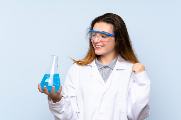 Young brunette girl over isolated blue wall with a scientific test tube and celebration