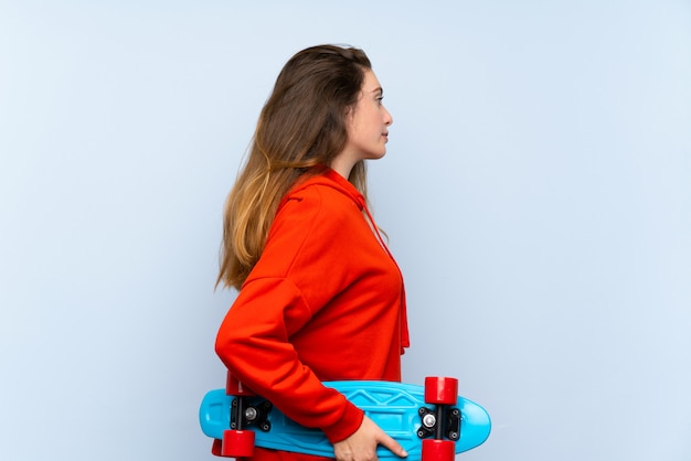 Young brunette girl over isolated blue wall with skate in back position