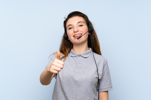 Young brunette girl over isolated blue wall working with headset with thumb up