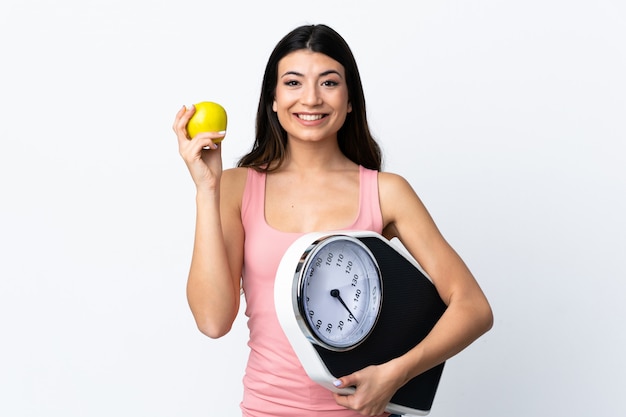 Young brunette girl over isolated white with weighing machine and with an apple