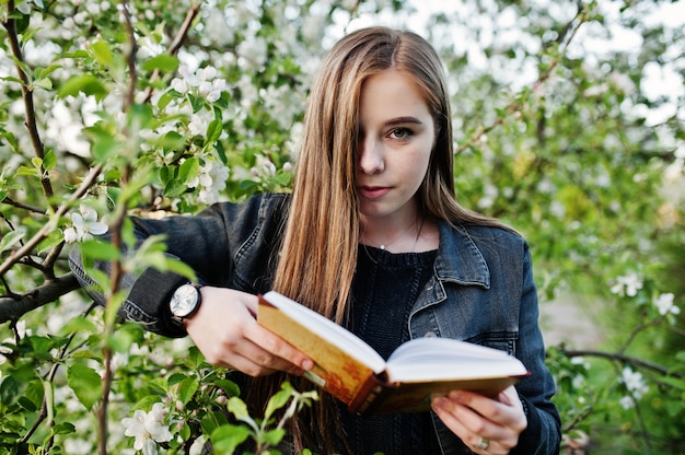 Young brunette girl at jeans against spring blossom tree read the book.