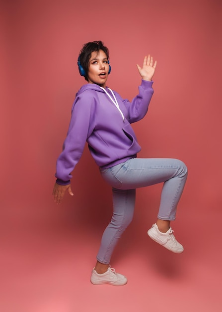 Young brunette girl in a lilac sweater and headphones poses in isolation on a pink background in the studio The concept of people's lifestyleListen to music with headphones dance studio portrait