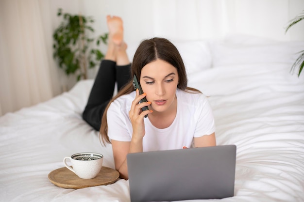 young brunette girl lying at home on the bed working at a laptop and talking on a mobile phone