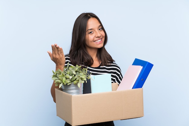 Young brunette girl making a move while picking up a box full of things inviting to come with hand. Happy that you came