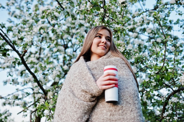 Young brunette girl on plaid against spring blossom tree and holding tea thermos at hands