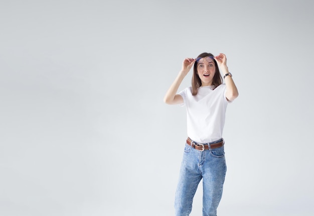 A young brunette girl raises her glasses in surprise and looks away in surprise on a white isolated