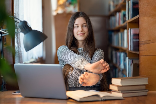 図書館のテーブルに座って手のストレッチをしている若いブルネットの女の子疲れた学生は、ハードな頭脳労働を勉強し、リラクゼーションのために一時停止するために多くの時間を費やしています