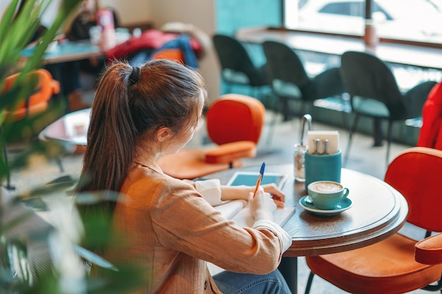 Young brunette girl student doing homework in notebook with coffee at the cafe