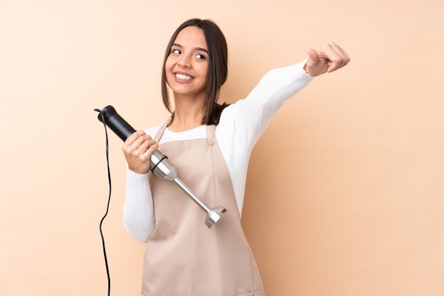 Young brunette girl using hand blender over isolated background giving a thumbs up gesture