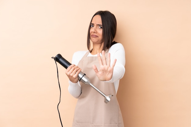 Young brunette girl using hand blender over isolated wall nervous stretching hands to the front