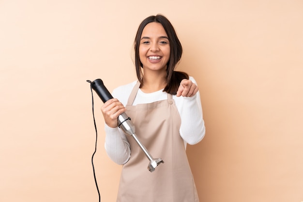 Young brunette girl using hand blender over isolated wall surprised and pointing front