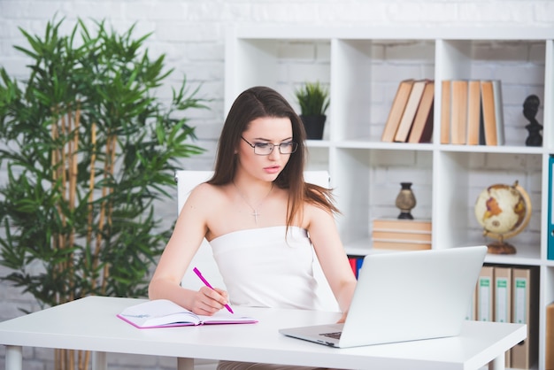 Una giovane ragazza bruna in un abito bianco è seduta in ufficio al tavolo e scrive note importanti sul suo taccuino. la donna lavora al computer portatile.