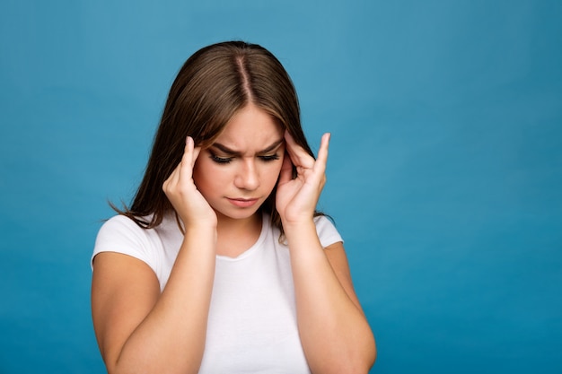 Young brunette girl in white t-shirt suffering from a headache, blue background