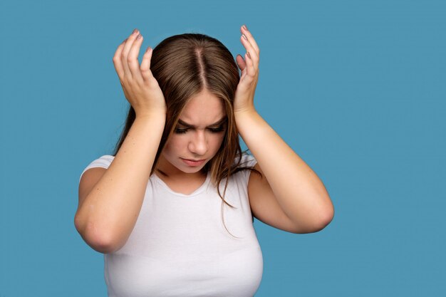 Young brunette girl in white t-shirt suffering from a headache, isolated on blue background