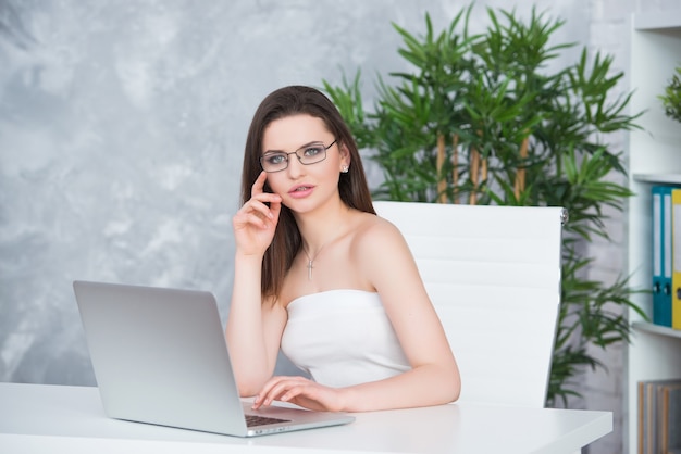 A young brunette girl with glasses in a white dress is sitting in the office at the table. Woman works at the laptop. Low vision or fashion accessory.