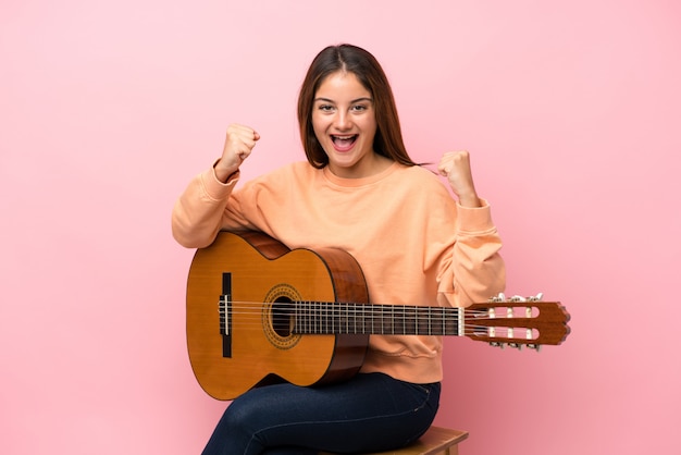 Giovane ragazza castana con la chitarra sopra il rosa isolato che celebra una vittoria