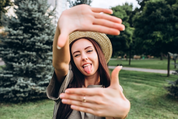 Фото Молодая брюнетка битник женщина берет selfie, показывая язык, смешное лицо позирует на открытом воздухе. портрет крупного плана девушки в стильной шляпе лета в парке.