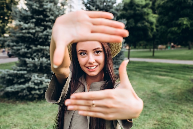 Selfieを取って、舌、屋外でポーズ変な顔を見せて若いブルネット流行に敏感な女性。公園でスタイリッシュな夏帽子の少女のポートレート、クローズアップ。